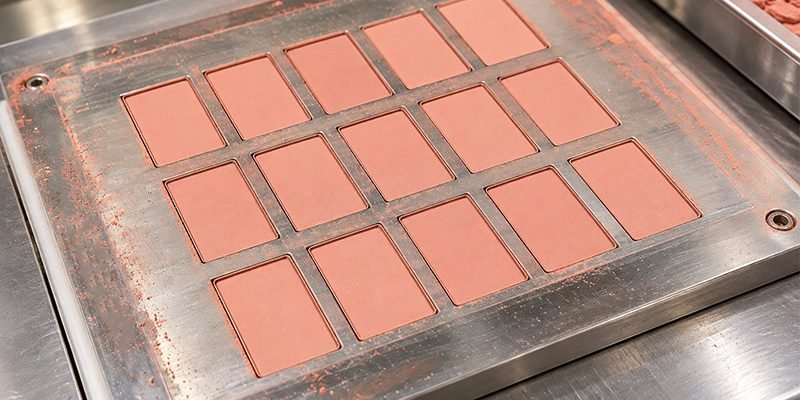 Pressed powder blush manufacturing with rectangular pink blush pans arranged in a metal production mold.
